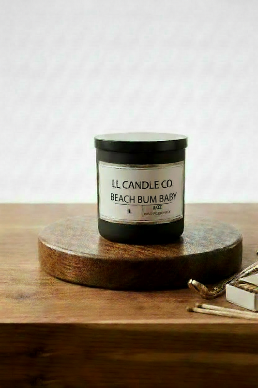 A jar candle labeled 'LL CANDLE CO. BEACH BUM BABY' sits on a round wooden coaster on a wooden table, with a matchbox and matches nearby against a light textured wall.