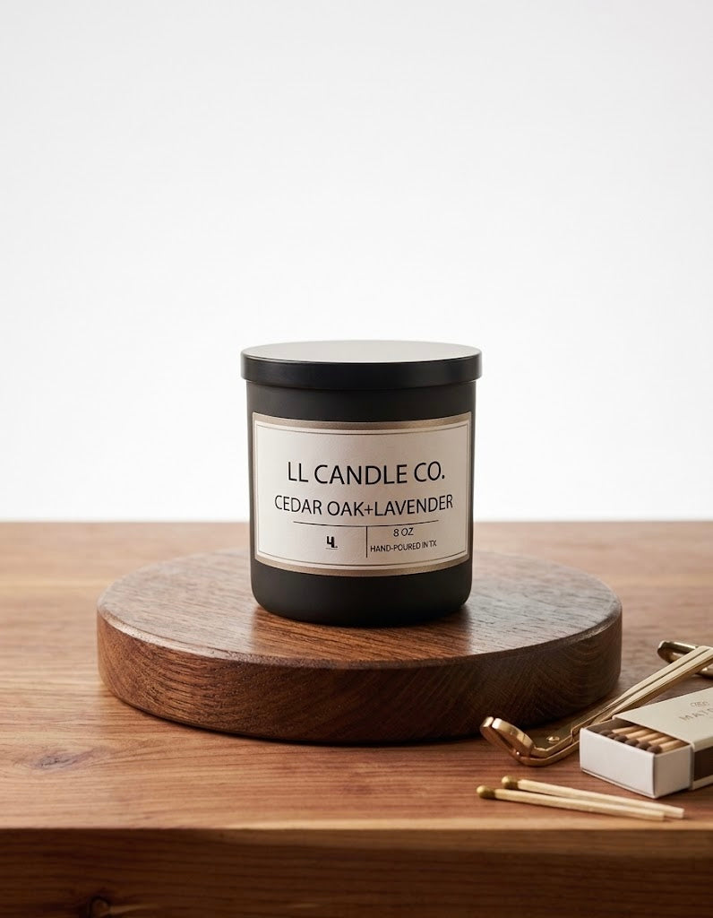 A black jar candle labeled 'LL CANDLE CO. CEDAR OAK + LAVENDER' sits on a round wooden pedestal on a wooden tabletop, with a matchbox and matches nearby against a clean white background.