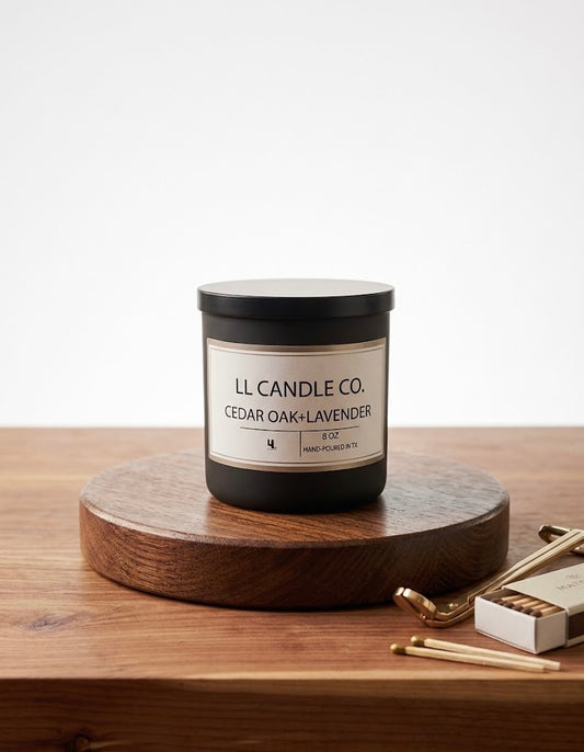 A black jar candle labeled 'LL CANDLE CO. CEDAR OAK + LAVENDER' sits on a round wooden pedestal on a wooden tabletop, with a matchbox and matches nearby against a clean white background.