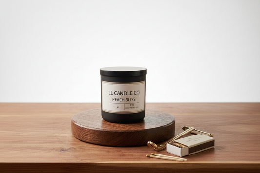 A black jar candle labeled 'LL CANDLE CO. PEACH BLISS' sits on a round wooden riser on a wooden tabletop, with a matchbox and matches beside it against a clean white background.