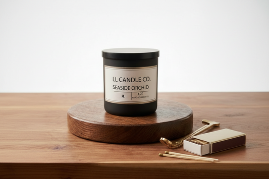 A black jar candle from LL Candle Co. labeled 'Seaside Orchid' sits on a round wooden coaster on a wooden table, with a gold-toned matchbox and matches beside it against a clean white background.