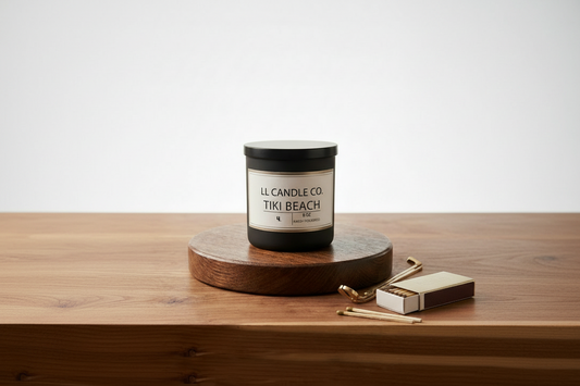 A black jar candle with a cream label reading 'LL CANDLE CO. TIKI BEACH' sits on a round wooden coaster on a wooden tabletop, with a matchbox and matches beside it against a clean white background.