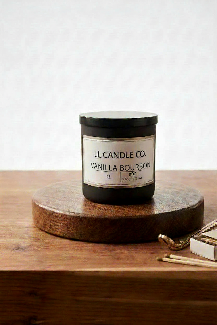 A jar candle labeled 'LL Candle Co. Vanilla Bourbon' sits on a round wooden coaster on a wooden table, with a few gold-toned matches nearby against a light textured wall.