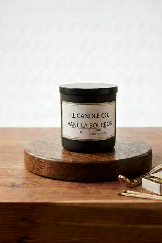 A jar candle labeled 'LL Candle Co. Vanilla Bourbon' sits on a round wooden coaster on a wooden table, with a few gold-toned matches nearby against a light textured wall.